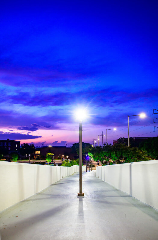 A UAB pedestrian bridge at night, an illuminated lamp in the middle, under a violet and pink sky. 