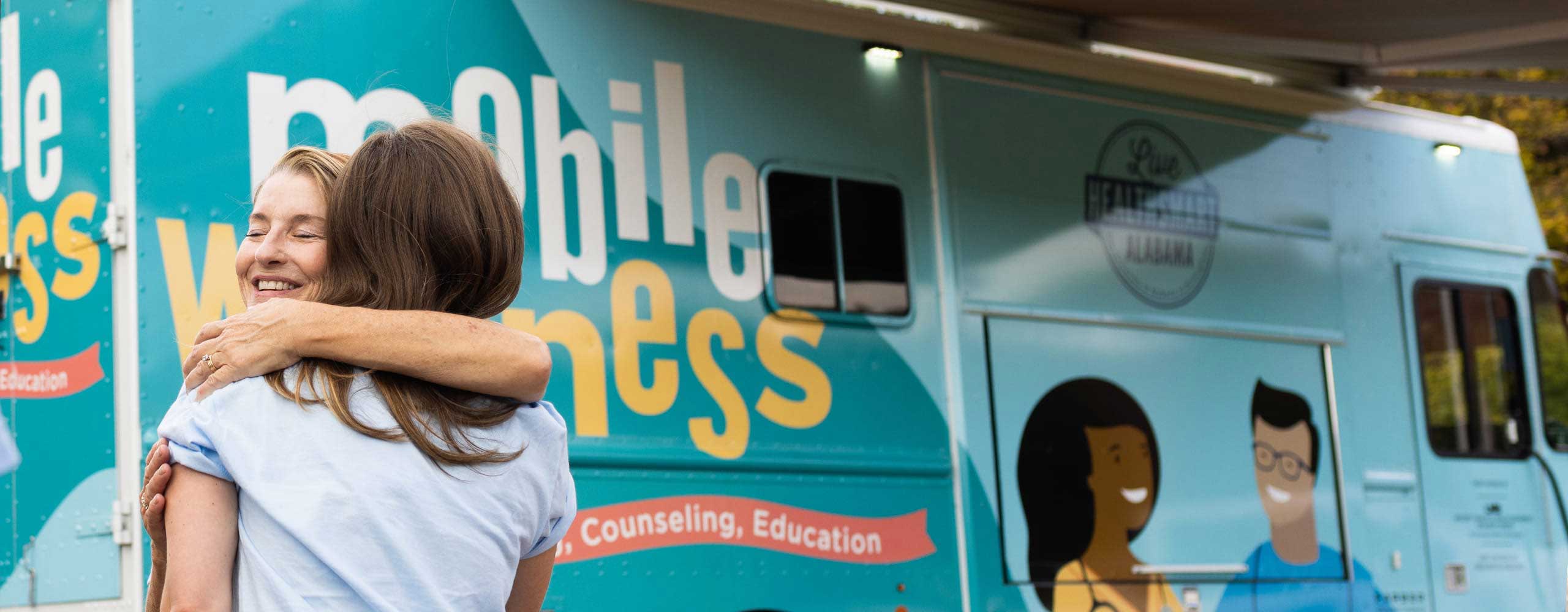 Two women hugging outside the light blue mobile wellness bus. 