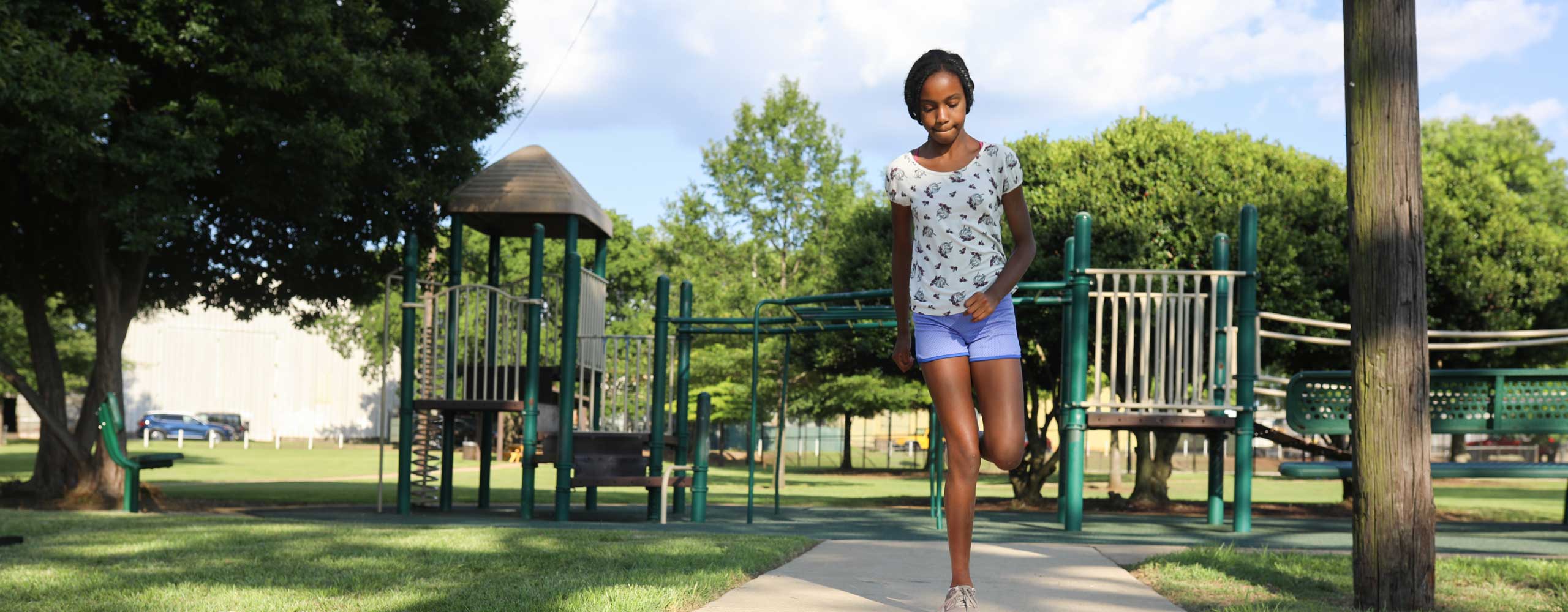 A young Black girl skips through a park, playground equipment in the background. 