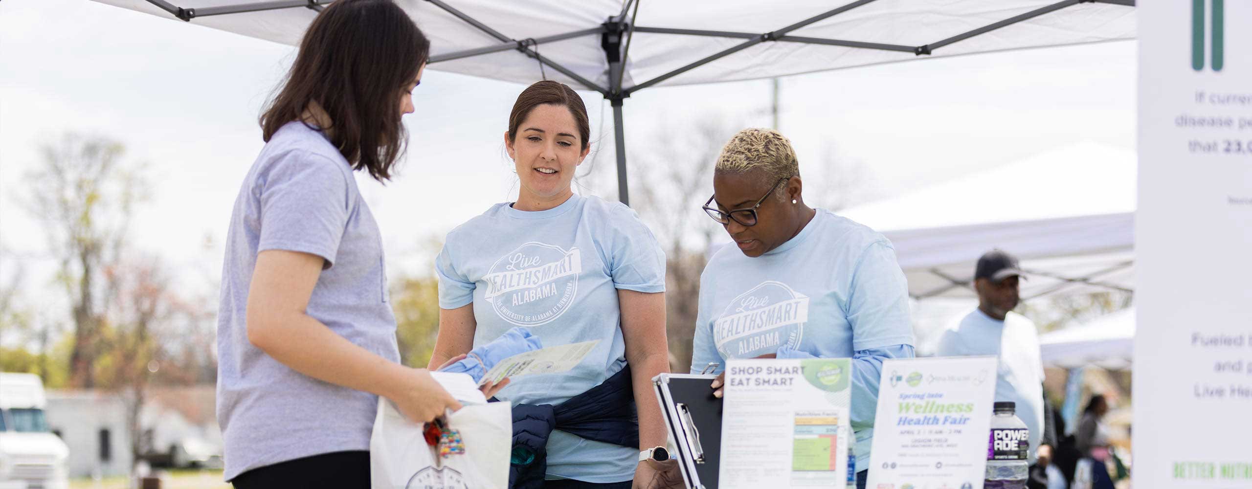 Young woman in a lavender tshirt talks to two women at an outdoor Live HealthSmart info table. 