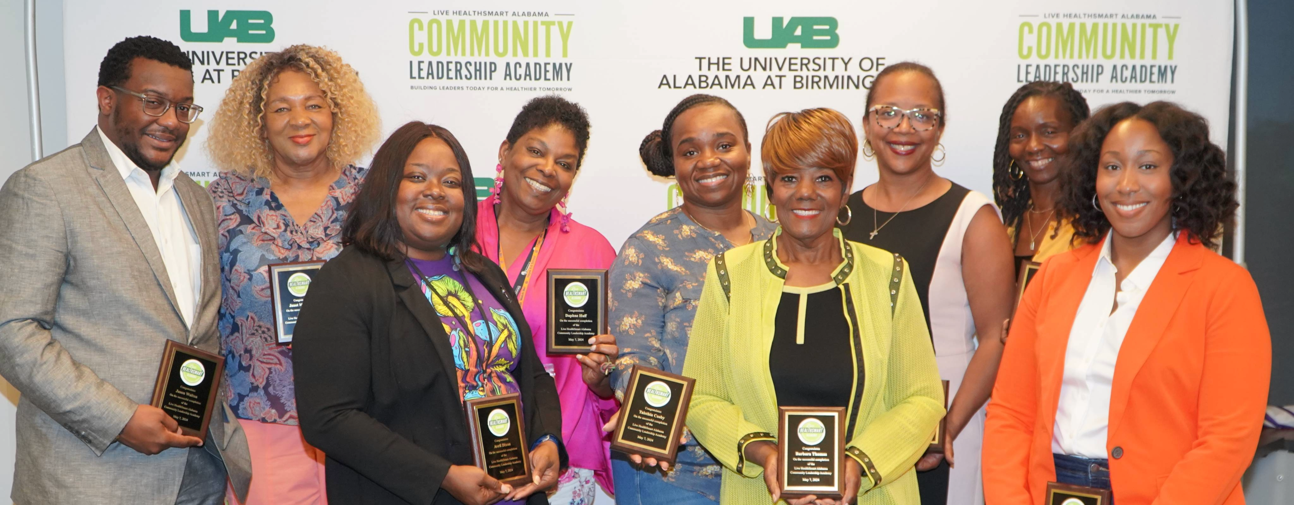 Group photo of people in a line with a man in a suit on the far left and eight women extending to the right with a woman in an orange blazer at the end, holding plaques marking their completion of the community leadership academy.