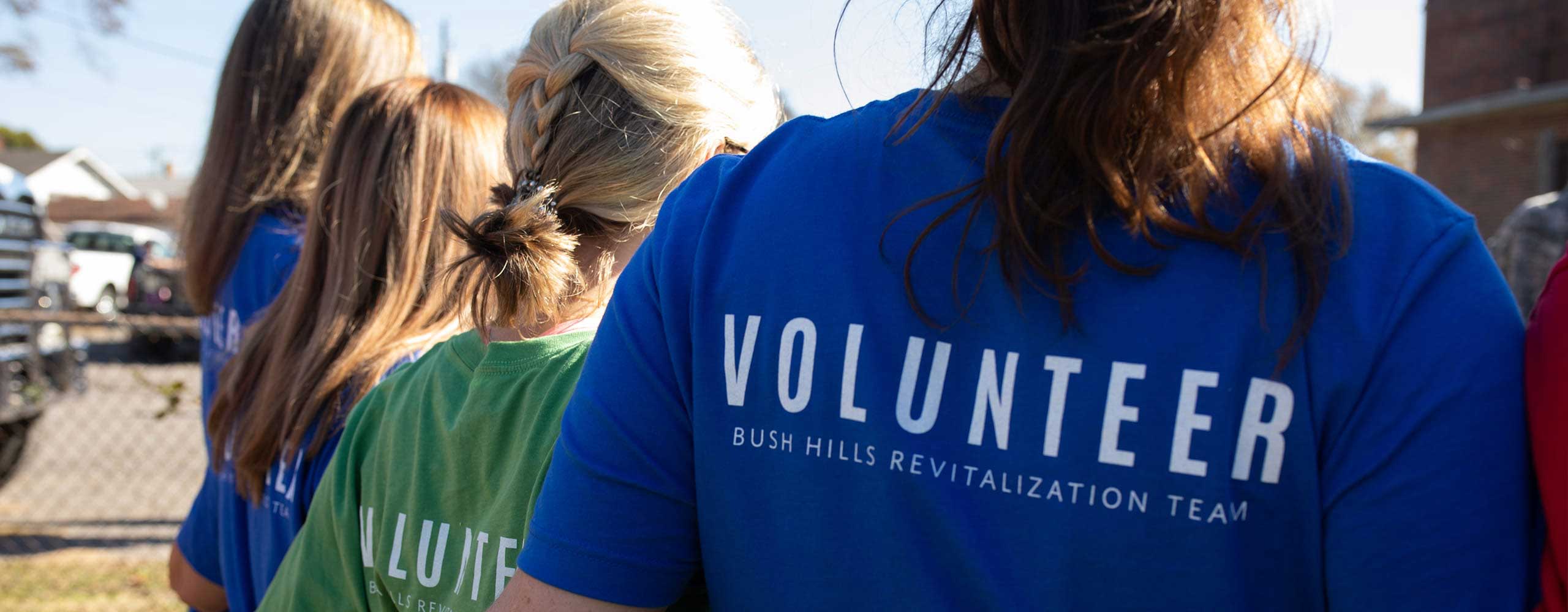 Close up on the backs of 4 people with long hair, the two closest to the camera in t-shirts reading volunteer.
