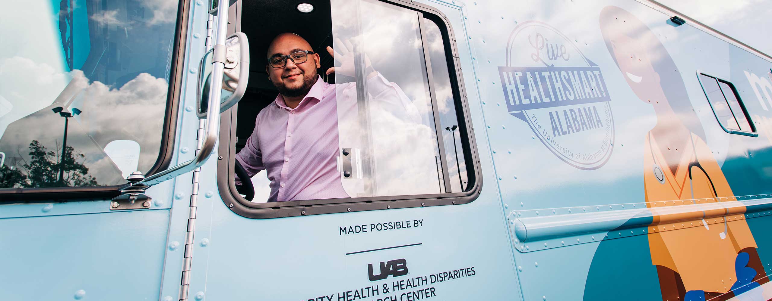 A man in glasses and a pink button-up waves from the driver's seat of the mobile wellness van.