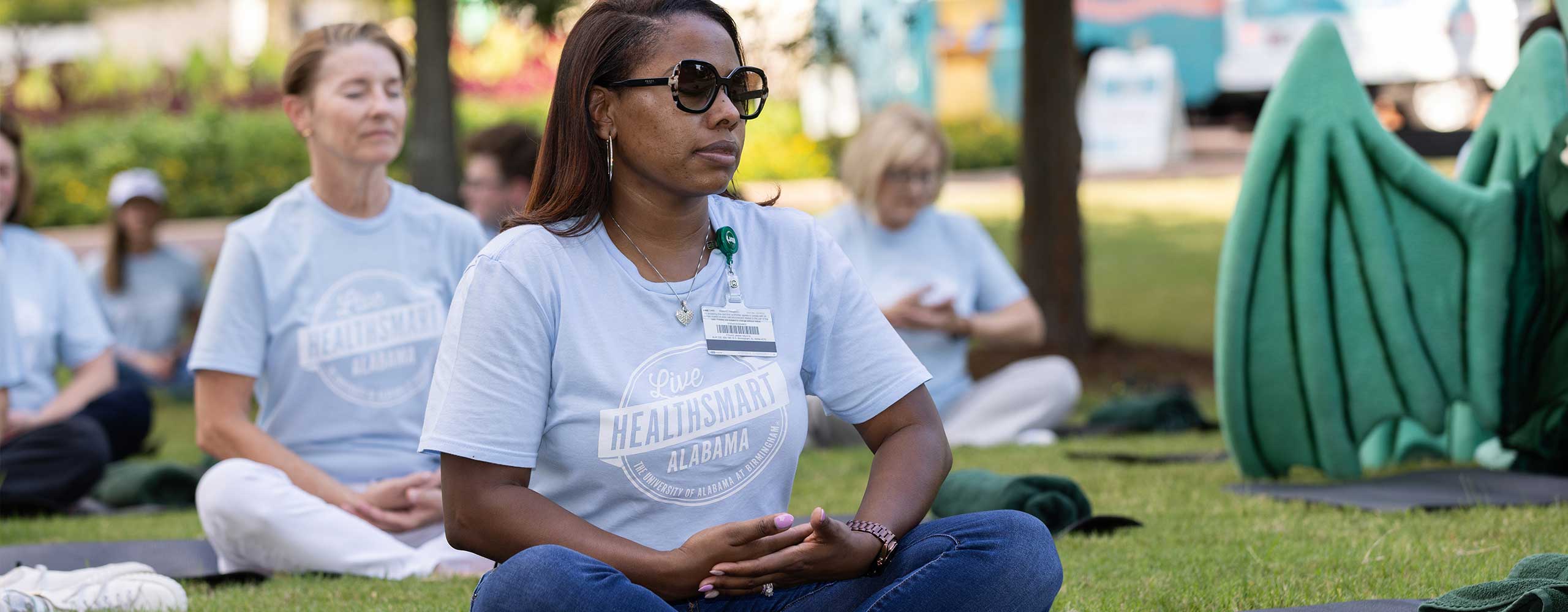 Women wearing Live HealthSmart shirts sit crosslegged on the grass meditating. 