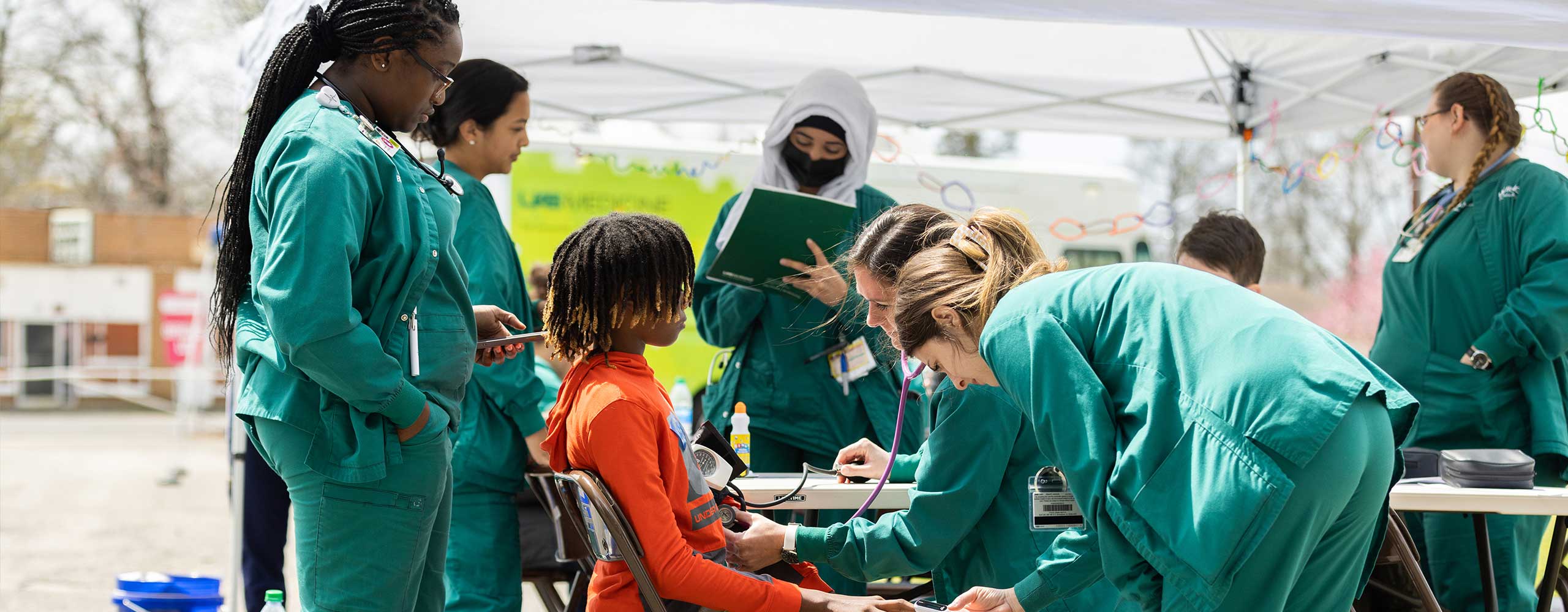 A young boy with locs being screened by several nurses in green scrubs in a tent. 