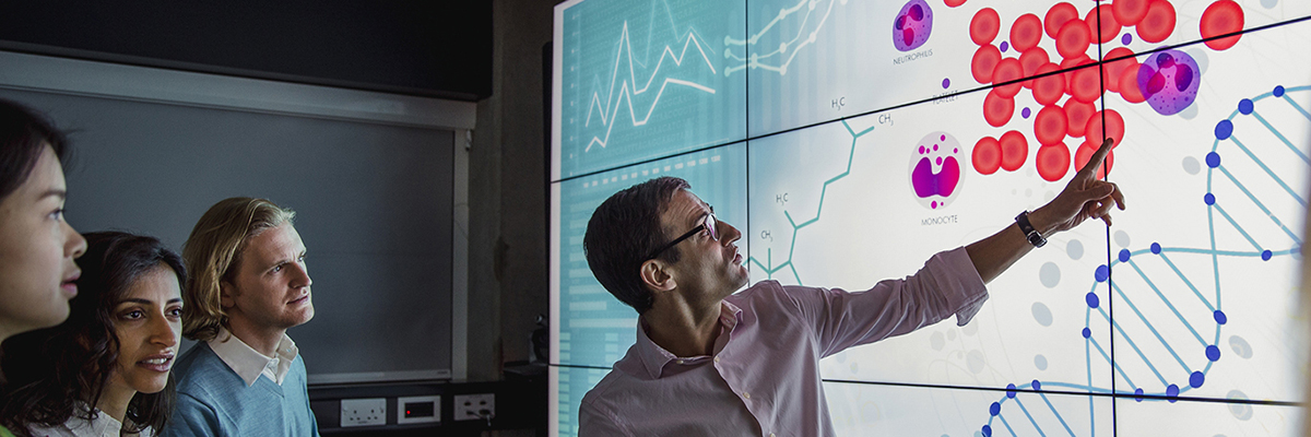 Group of business professionals in a dark room standing in front of a large data display screen with information.