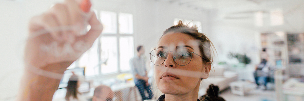 Photo of a young woman writing on a transparent wipe board and thinking of a solution for her work-related problems