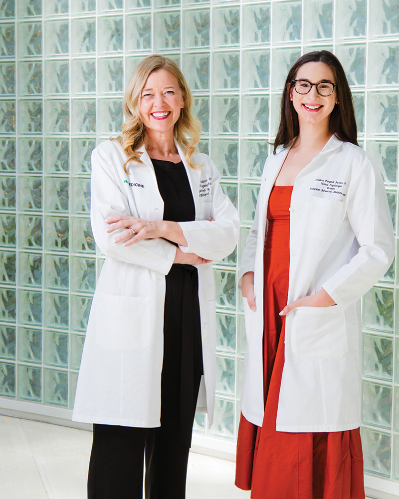 Kristine Lokken, Ph.D., and Christina Pierpaoli Parker, Ph.D., of the UAB Department of Psychiatry and Behavioral Neurobiology pose for portraits in their white coats. They stand in front of a wall of glass bricks in the lobby of the Kirklin Clinic. 