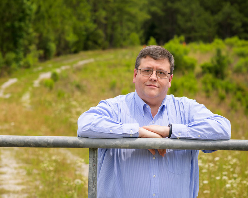 Dr. Drake Lavender poses with his arms resting on a metal gate in a rural scene. A dirt road curves into the distance behind him. 