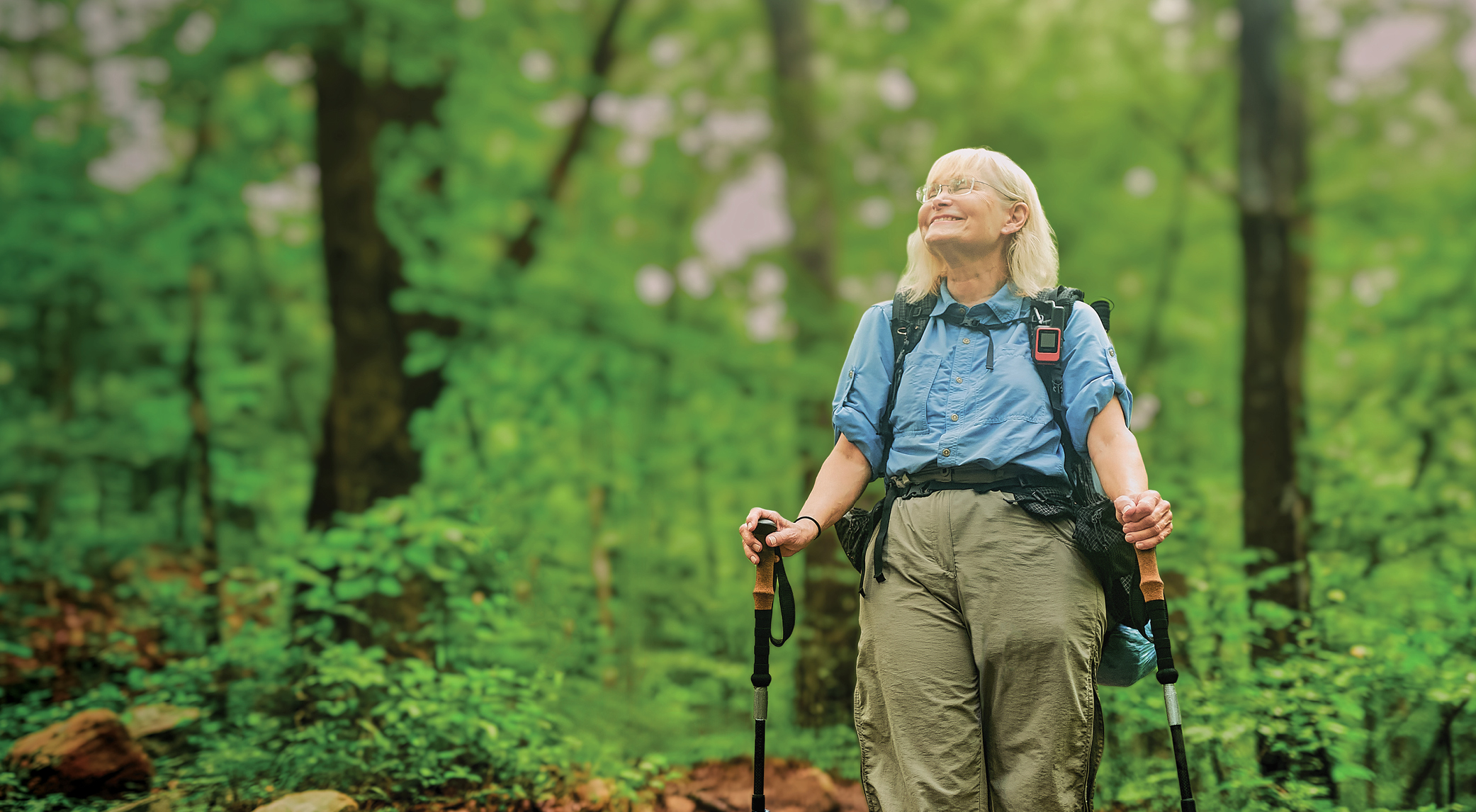 Cindy Louderback flashes a skyward smile while hiking a lush green trail at Red Mountain. She wears a blue shirt and khaki pants and is using hiking poles.