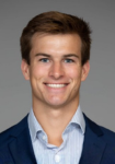 Professional headshot of a man wearing a navy suit jacket with a light blue shirt  in front of a grey backdrop