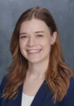 Professional headshot of a woman wearing a navy suit jacket with a white shirt  in front of a blue backdrop