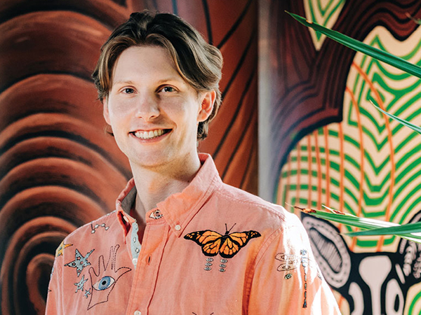 A portrait of medical student Matthew Kiszla. He wears a salmon shirt with hand-embroidery and is pictured against a brightly colored mural background.
