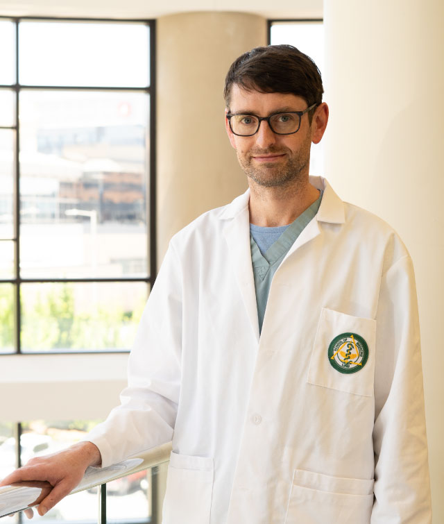 UAB neonatologist Dr. Colm Travers wearing a white coat stands in a UAB Hospital building near large windows, looking toward the camera.