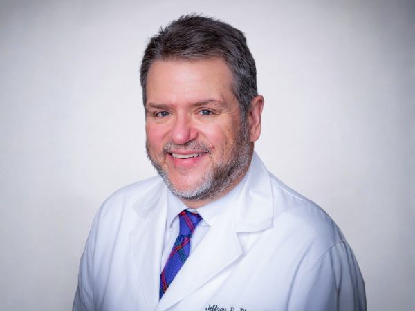 Professional headshot of a smiling man with short dark-and-gray hair and a trimmed beard, wearing a white medical coat, white shirt, and blue patterned tie against a light gray background.