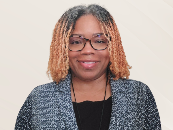 Headshot of a person wearing glasses, a black top, and a textured black-and-white blazer. The person has shoulder-length curly hair with lighter-colored ends and is facing the camera against a plain light background. 