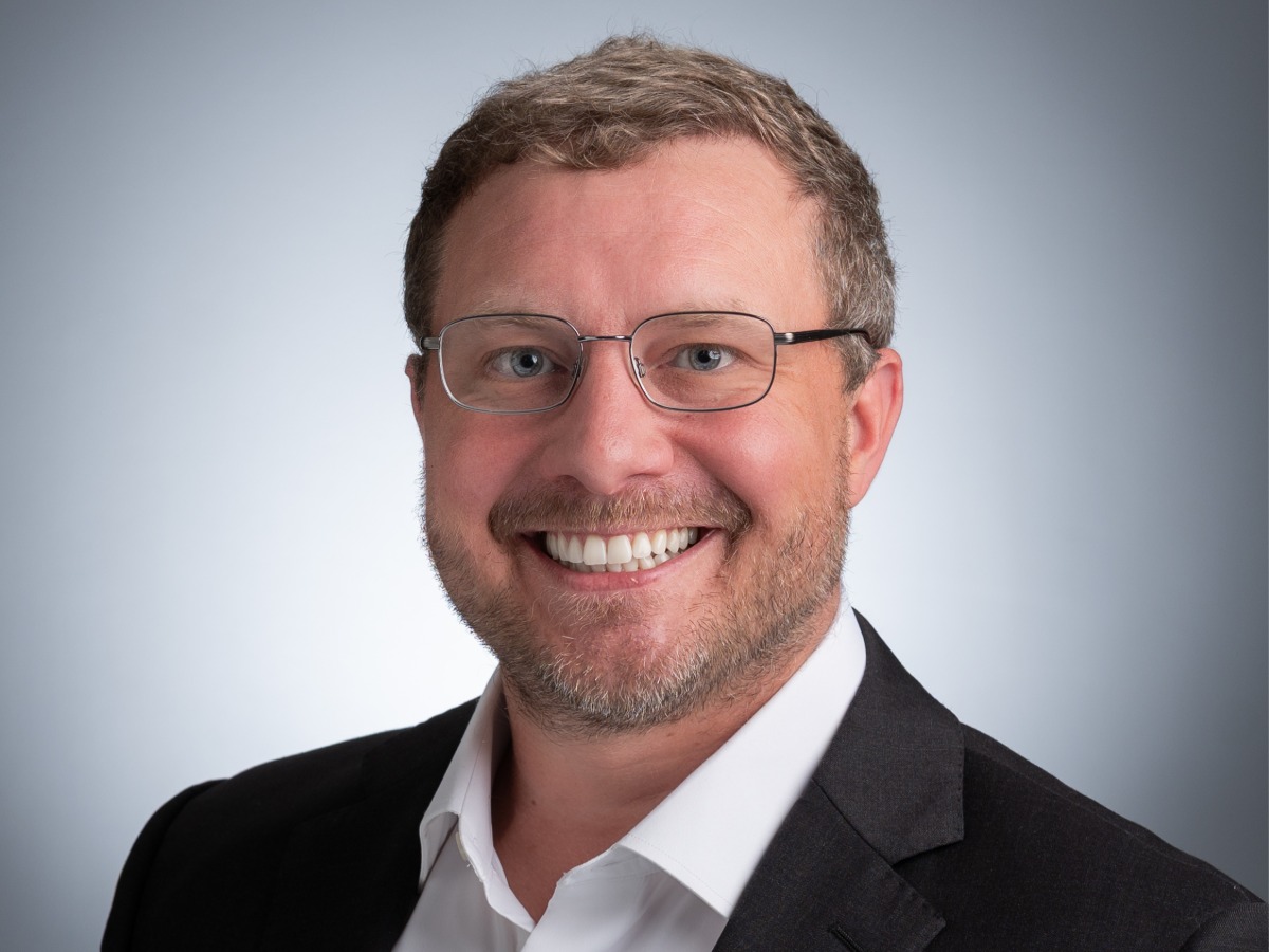Photo of Tim Johnson, MD, smiling and wearing glasses, a black blazer, and white buttoned shirt against a grey background.