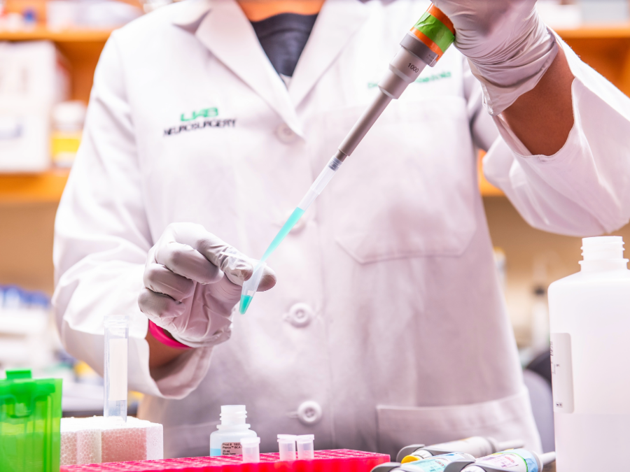 A person wearing a white lab coat and gloves uses a pipette to transfer a blue liquid into a small test tube on a laboratory bench. Various lab containers, racks, and bottles are visible in the background.