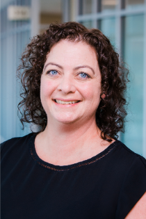 Headshot of a person with curly shoulder-length hair, wearing a dark top, in front of a softly blurred glass background.