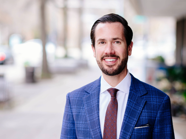 Portrait of a person in a blue suit and tie, with trimmed facial hair, standing outdoors with a blurred background.