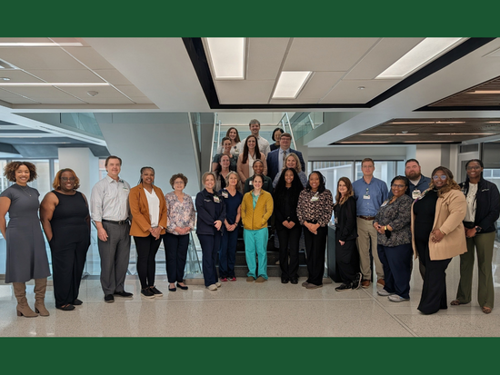 A large group of about 26 healthcare professionals stand together for a group photo inside a bright, academic building. They are arranged in two rows—some on a staircase and others standing in front—smiling and facing the camera. Most are wearing business or clinical attire, including scrubs, lab coats, and name badges. The space features glass railings, wide hallways, and overhead lighting panels, with an illuminated red exit sign visible in the background.