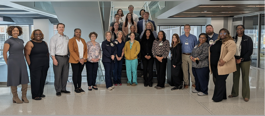 A large group of about 26 healthcare professionals stand together for a group photo inside a bright, academic building. They are arranged in two rows—some on a staircase and others standing in front—smiling and facing the camera. Most are wearing business or clinical attire, including scrubs, lab coats, and name badges. The space features glass railings, wide hallways, and overhead lighting panels, with an illuminated red exit sign visible in the background.