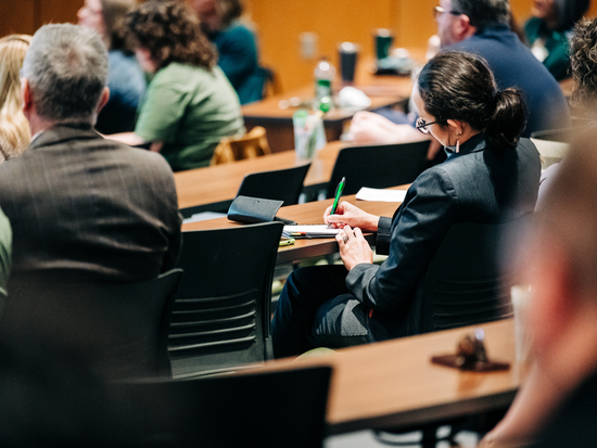 Attendees seated in a lecture hall during a presentation a woman in business attire takes notes with a green pen while others listen in rows of tiered seating