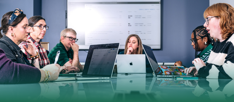 Group of professionals seated around a conference table collaborating with laptops and notes while looking at a presentation screen during a team meeting