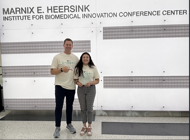 Two individuals stand smiling in front of a white feature wall inside the Marnix E. Heersink Institute for Biomedical Innovation Conference Center. Both are wearing matching light-colored event T-shirts with a green logo and holding small objects in their hands. The wall behind them displays the institute’s name in large lettering with horizontal textured design elements. The setting appears bright and modern with a polished floor and open space.
