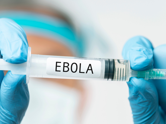 Close-up of gloved hands holding a syringe labeled “EBOLA,” with the healthcare worker in the background intentionally out of focus. The image represents Ebola-related medical research, vaccination, treatment, or infectious disease response.