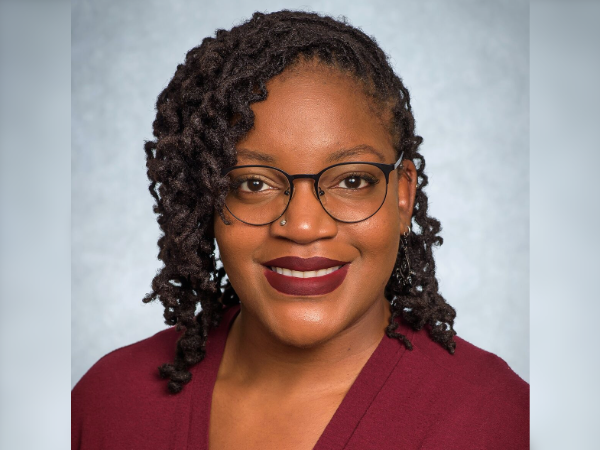 Professional headshot of an individual wearing glasses and a burgundy top against a light gray background.