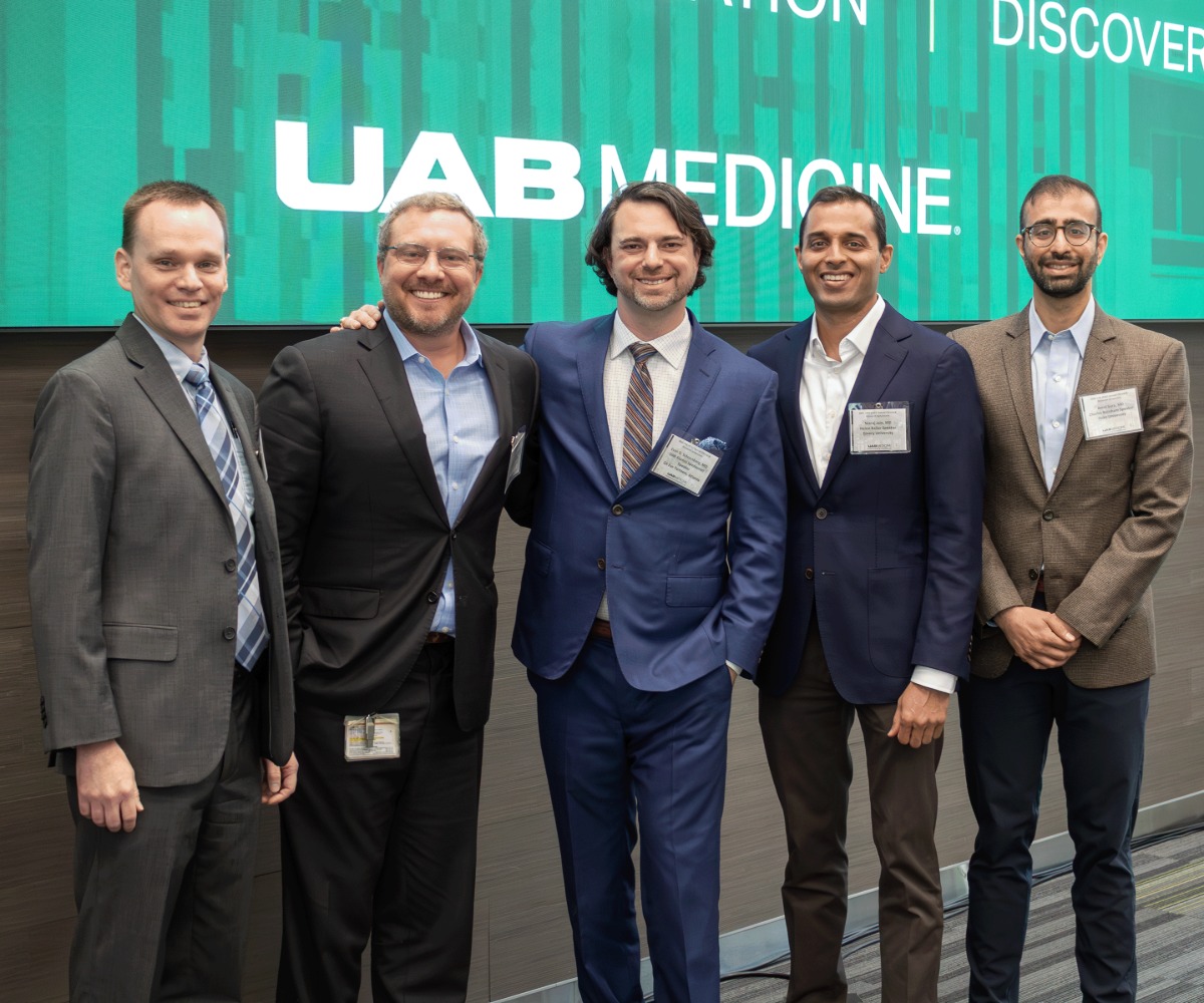 Five attendees pose together in front of a UAB Medicine conference display at a symposium, wearing business attire.