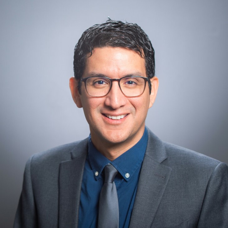 Javier Neyra headshot - man with dark hair and glasses wearing a blue button up shirt, tie and blazer jacket