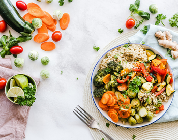 Bowl of cooked grains, lettuce, and mixed vegetables on a table surrounded by fresh vegetables.