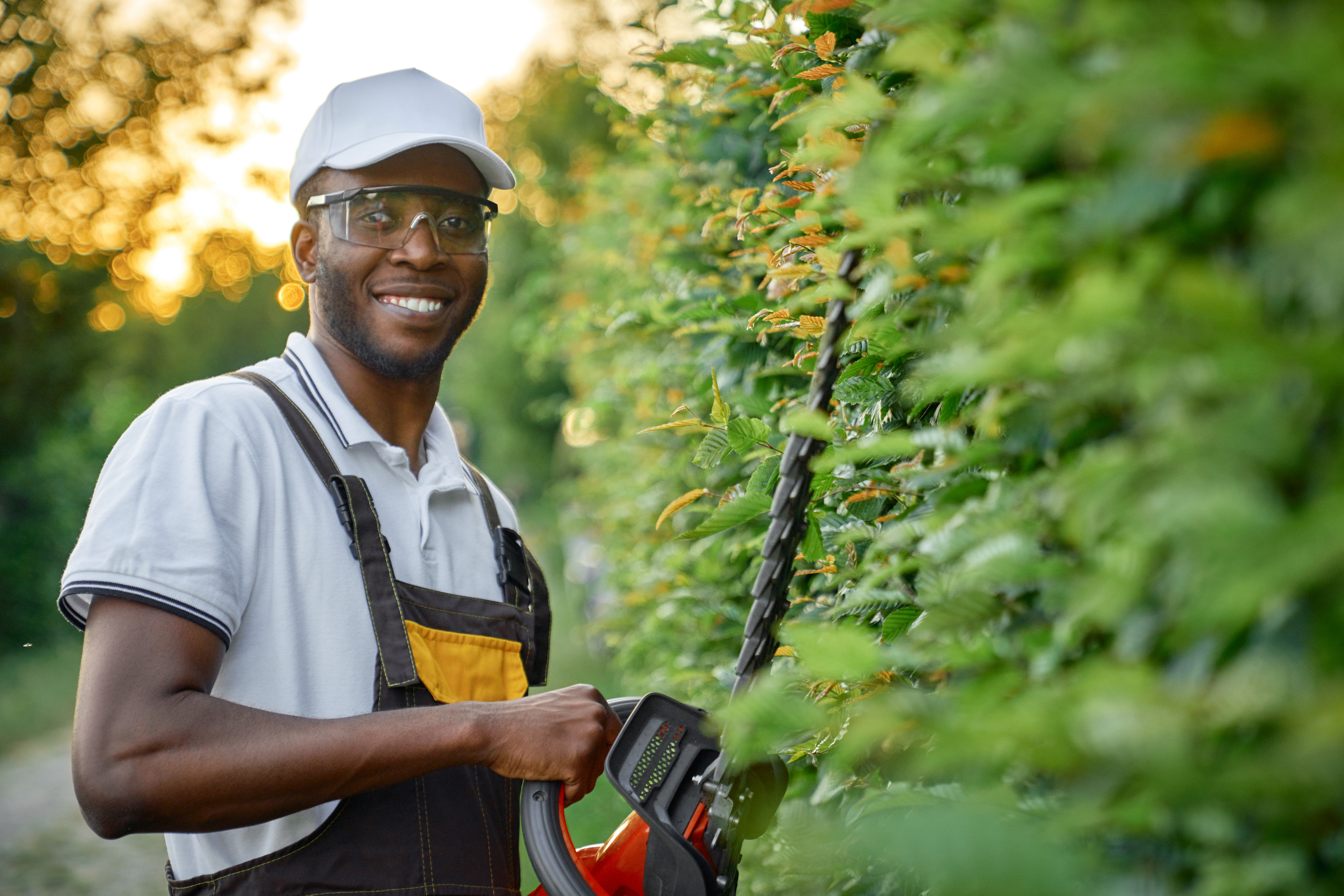 Portrait of handsome afro american guy in protective glasses and white summer hat smiling and looking at camera while standing near green decorative hedge with petrol trimmer. Concept of gardening.