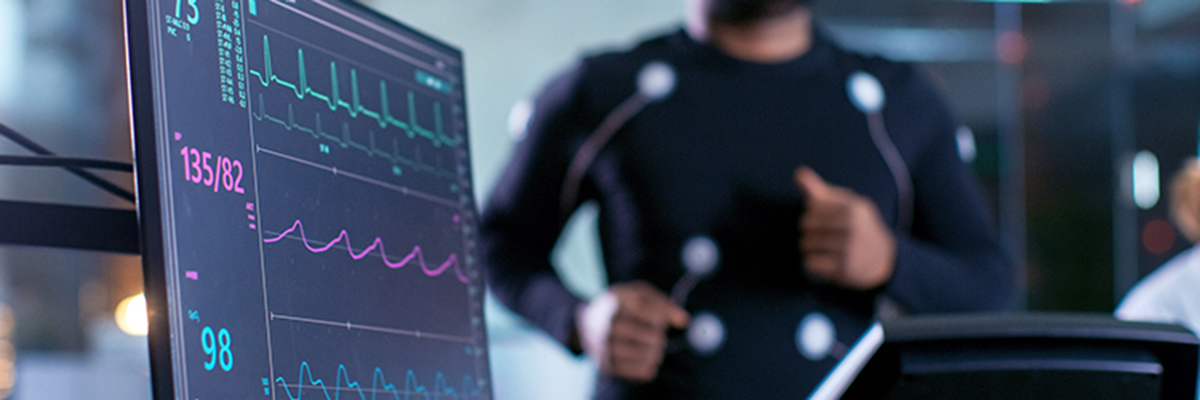 Close-up Shot of a Monitor With EKG Data. Male Athlete Runs on a Treadmill with Electrodes Attached to His Body while Sport Scientist Holds Tablet and Supervises EKG Status in the Background.