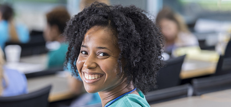 A young female medical student sits at a desk in a lecture hall.  She pauses from writing and turns to smile for the camera.