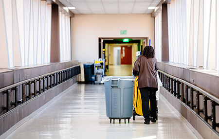 Back, a black female Environmental Services Specialist - Hospital is wearing a PPE (Personal Protective Equipment) face mask while walking behind janitorial cart in interior hospital crosswalk during the COVID-19 (Coronavirus Disease) pandemic, May 2020.