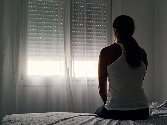 Rear view of an unrecognizable abused woman sitting on her bed looking out the window. Concept of gender violence, domestic violence and depression.