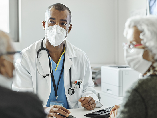 Doctor discussing with elderly couple during COVID-19 outbreak. Male healthcare worker is explaining senior man and woman while sitting at desk in clinic. They are in protective face masks.