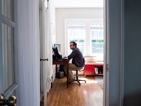 From side, Jordan Lingo (Graduate Student, Microbiology) is working at a computer on a desk in a room at his home, Apri 2020.