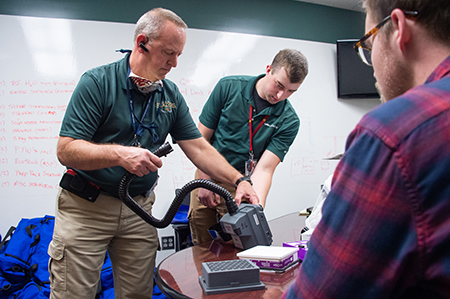 Bill Mayfield and Matthew Mayfield put the 3d printed filter reservoir into the vetinlator for a fit test as Joseph Moore looks on