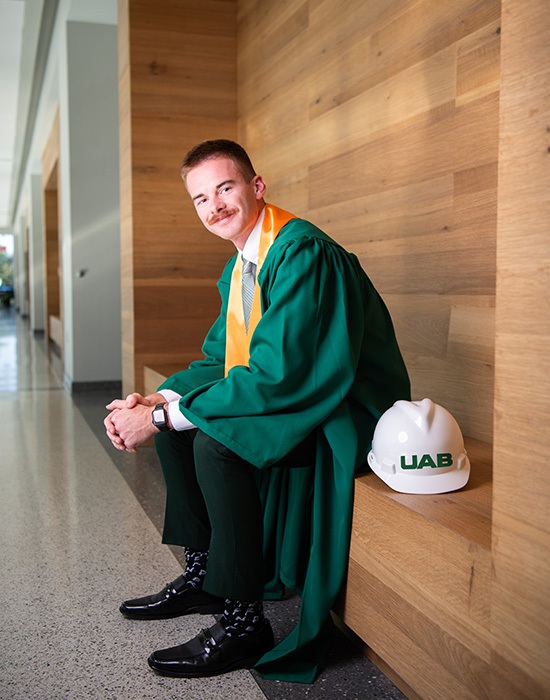  Jonah Ross in his graduation gown sitting with a UAB hard hat sitting next to him. 