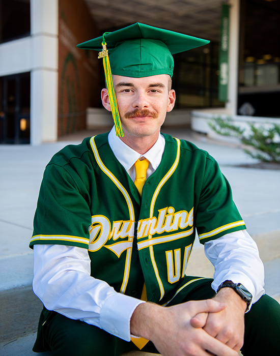  Jonah Ross sitting on steps outside of the Science and Engineering Complex with a drum line shirt and a UAB cap. 