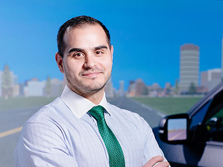 Environmental headshot of Dr. Benjamin McManus, PhD (Postdoctoral Fellow, Psychology) standing in front of the Driving Simulator in the TRIP (Translational Research for Injury Prevention) Laboratory, 2020.