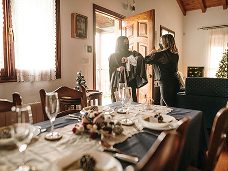 Coming over for Christmas dinner during the COVID-19 pandemic. Two women greet each other at the entrance of a house, with a Christmas decorated table for dinner