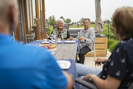 two senior couples sitting together on a long table outdoors in garden enjoying afternoon coffee and sweet snacks, both sitting on one end to keep social distance in times of loosend coronavirus curfew