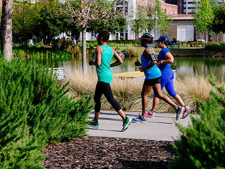 From side, three black women are jogging on paved trail in Railroad Park, 2019.