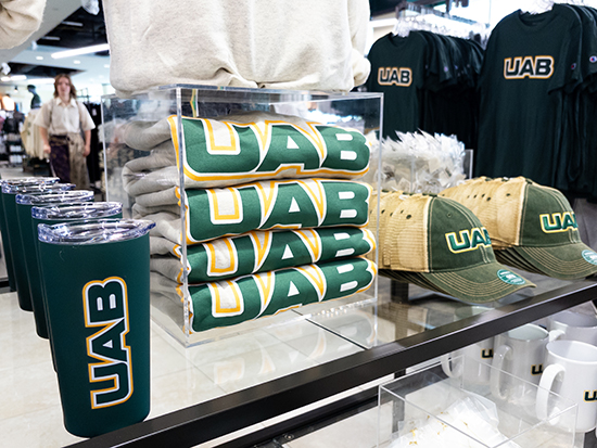 Interior of UAB University Bookstore showing water bottles, caps, and various items of clothing with the new University logo on display on table and racks.