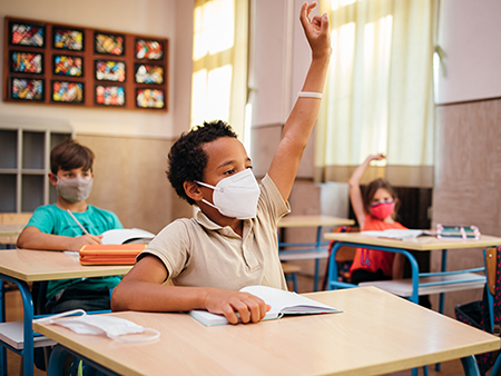 African-American schoolboy with protective mask is sitting at a desk in the classroom with raised hand in desire to answer the question.